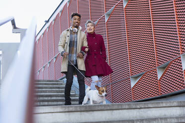 Lächelndes junges Paar, das mit seinem Hund eine moderne Stadttreppe hinuntergeht - CAIF27246