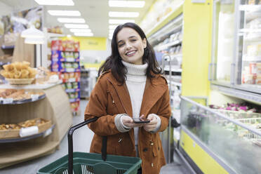 Portrait lächelnde, selbstbewusste Frau mit Smartphone beim Einkaufen im Supermarkt - CAIF27299