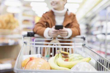 Frau mit Smartphone schiebt Einkaufswagen im Supermarkt - CAIF27301