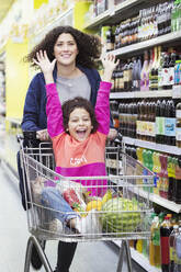 Mutter schiebt ihre aufgeregte Tochter in einem Einkaufswagen durch einen Supermarktgang - CAIF27304