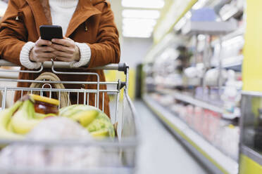 Frau mit Smartphone schiebt Einkaufswagen im Supermarkt - CAIF27306