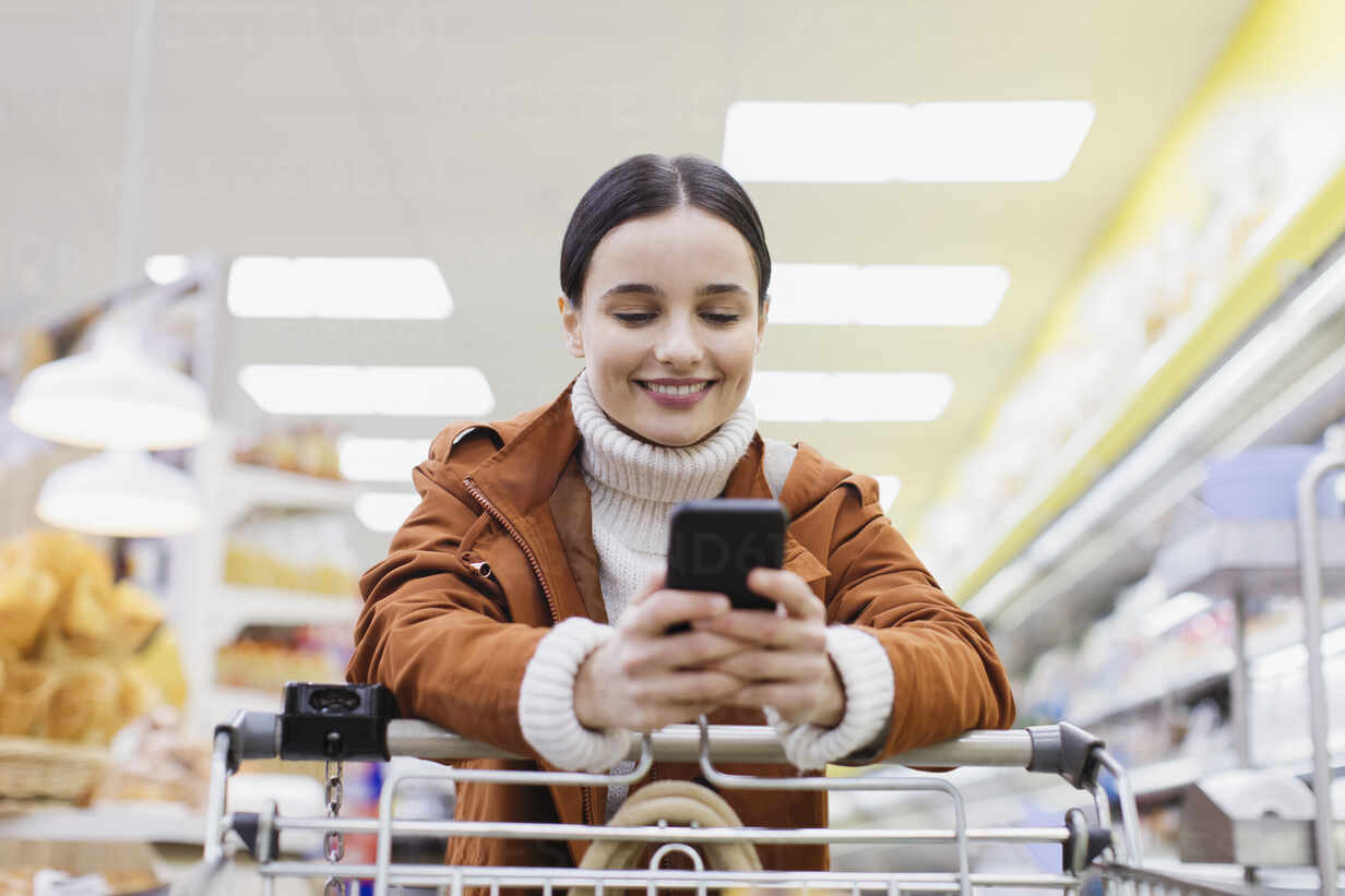 Woman with smart phone pushing shopping cart in supermarket stock photo