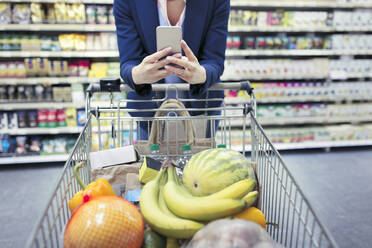 Frau mit Smartphone schiebt Einkaufswagen im Supermarkt - CAIF27334