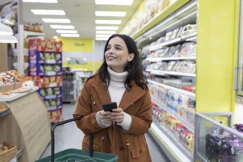 Lächelnde junge Frau mit Smartphone beim Lebensmitteleinkauf im Supermarkt - CAIF27351