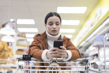 Junge Frau mit Smartphone beim Einkaufen im Lebensmittelladen - CAIF27354