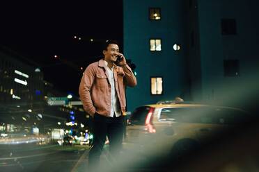 Smiling man talking through smart phone while standing in illuminated city at night - MASF17939