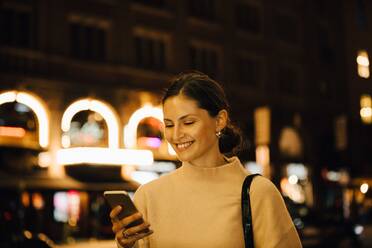 Lächelnde Frau, die nachts in der Stadt steht und eine SMS in ihr Handy spricht - MASF17954