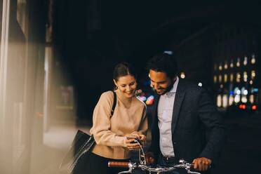 Lächelnde Frau zeigt einem männlichen Freund mit Fahrrad ihr Smartphone, während sie nachts in der Stadt steht - MASF17957