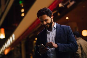 Niedriger Blickwinkel eines lächelnden Mannes, der ein Smartphone mit In-Ear-Kopfhörern benutzt, während er in einer nächtlichen Stadt steht - MASF17960