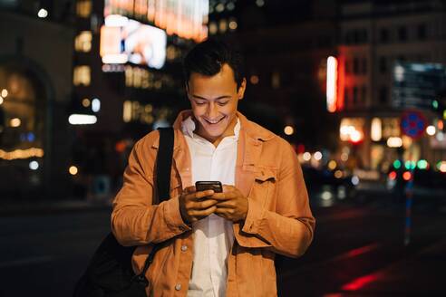 Lächelnder junger Mann, der eine SMS in sein Handy spricht, während er in einer beleuchteten Stadt bei Nacht steht - MASF17965