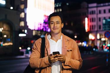 Portrait of smiling man using mobile phone while standing in illuminated city at night - MASF17966