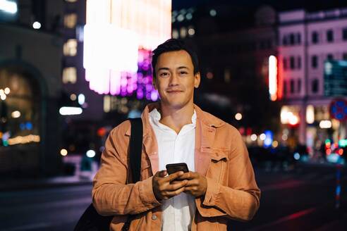 Porträt eines lächelnden Mannes, der ein Mobiltelefon benutzt, während er in einer beleuchteten Stadt bei Nacht steht - MASF17966