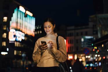 Smiling woman texting through smart phone in city at night - MASF17968