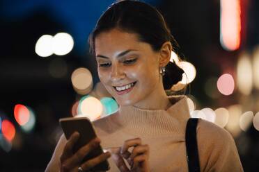 Lächelnde junge Frau, die in der nächtlichen Stadt eine SMS über ihr Smartphone schreibt - MASF17972