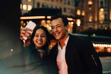 Lächelnder Mann und Frau nehmen Selfie durch Telefon, während in der Stadt in der Nacht stehen - MASF17976