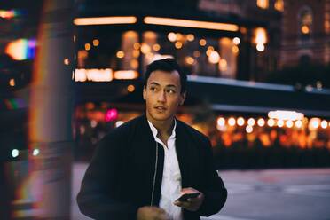 Young man with phone looking away while standing in illuminated city at night - MASF17984