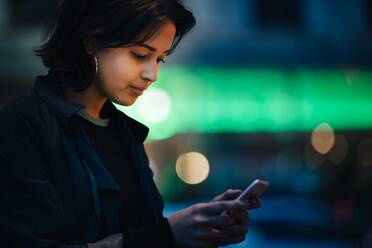 Junge Frau, die nachts in der Stadt steht und eine SMS auf ihrem Mobiltelefon schreibt - MASF17989