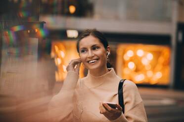 Young woman using in-era headphones while standing in city - MASF17996