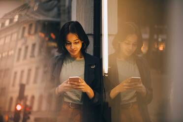 Junge Frau, die eine SMS in ihr Telefon spricht, während sie an einem Glasfenster steht - MASF18003