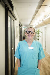 Porträt einer reifen Mitarbeiterin des Gesundheitswesens mit den Händen in den Taschen, die in einem Krankenhauskorridor steht - MASF18040