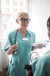 Smiling mature pediatrician gesturing while talking to female healthcare worker in clinic - MASF18044