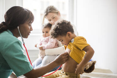 Seitenansicht einer Ärztin, die den Herzschlag eines Jungen mit einem Stethoskop untersucht, während Mutter und Tochter in der Klinik sitzen - MASF18050
