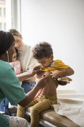 Ärztin, die den Herzschlag eines Jungen mit einem Stethoskop untersucht, während Mutter und Tochter in der Klinik sitzen - MASF18052