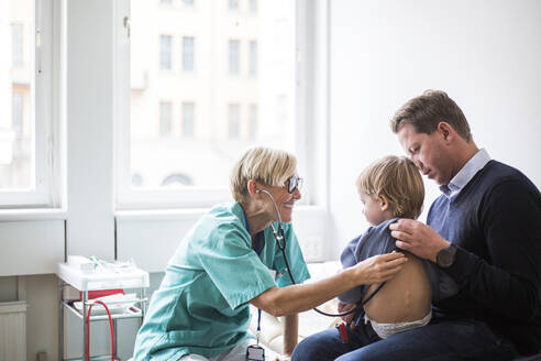 Lächelnde Ärztin, die den Rücken eines Jungen mit einem Stethoskop untersucht, während er neben seinem Vater im Krankenhaus sitzt - MASF18056