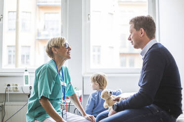 Seitenansicht einer reifen Ärztin im Gespräch mit einem Mann, während der Sohn im Hintergrund in einer medizinischen Klinik steht - MASF18060