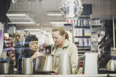 Saleswoman showing kettle to female customer in store seen through glass window - MASF18074
