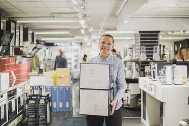 Portrait of smiling mature female owner with boxes standing in electronics store - MASF18101