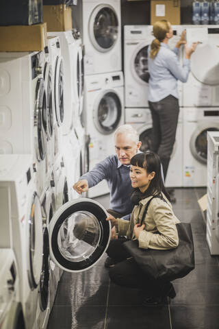 High angle view of mature owner pointing to washing machine while female customer crouching in electronics store stock photo