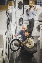 High angle view of mature owner pointing to washing machine while female customer crouching in electronics store - MASF18112