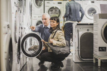 Mature owner pointing to washing machine while female customer crouching in electronics store - MASF18114