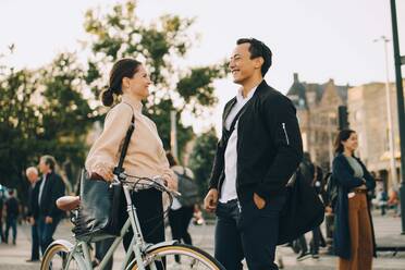 Smiling woman with bicycle looking at male friend while standing in city - MASF18151