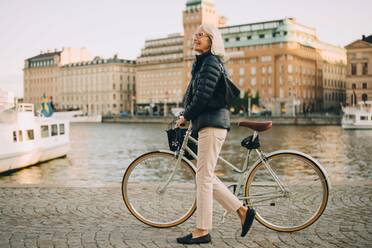 Seitenansicht einer lächelnden älteren Frau mit Fahrrad, die beim Überqueren eines Flusses in der Stadt nach oben schaut - MASF18155