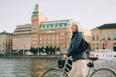 Seitenansicht einer lächelnden älteren Frau mit Fahrrad beim Überqueren eines Flusses in der Stadt - MASF18157