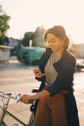 Young woman surfing net while standing with bicycle in city - MASF18201