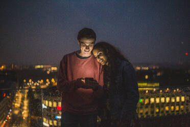 Junger Mann zeigt einer Freundin sein Smartphone während eines geselligen Beisammenseins auf einer Terrasse bei Nacht - MASF18205