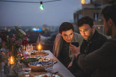 Mann zeigt männlichen Freunden sein Smartphone, während er bei einem geselligen Beisammensein auf der Terrasse sitzt - MASF18209