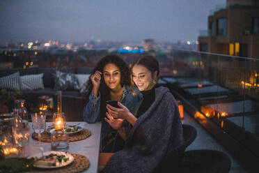 Lächelnde Frau zeigt einer Freundin ihr Smartphone während eines geselligen Beisammenseins auf der Terrasse eines Gebäudes - MASF18210