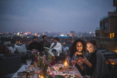Lächelnde Frau, die einer Frau ihr Smartphone zeigt, während im Hintergrund Freunde bei einem geselligen Beisammensein auf der Terrasse eines Gebäudes sitzen - MASF18211