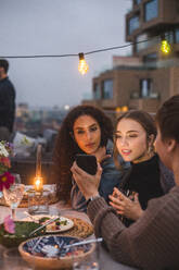 Frau zeigt Smartphone an Freundinnen bei geselligem Beisammensein auf der Terrasse eines Gebäudes - MASF18212