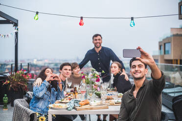 Lächelnder Mann, der ein Selfie mit seinem Smartphone macht, während er sich mit Freunden auf der Terrasse eines Gebäudes trifft - MASF18219