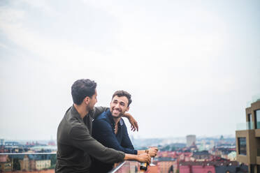 Happy man looking at male friend with drink while leaning over glass railing on rooftop against sky - MASF18245