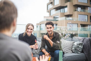 Lächelnder Mann und lächelnde Frau schauen ihre Freundin an, während sie auf dem Sofa bei einer Party auf der Terrasse eines Gebäudes sitzen - MASF18251