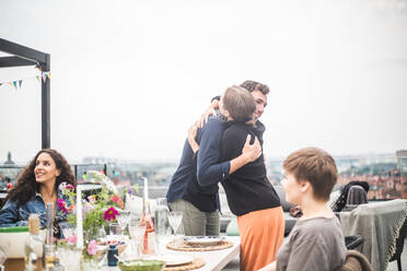 Young male and female friends embracing while standing by table on terrace - MASF18264