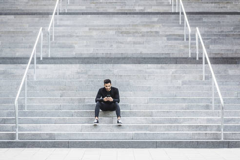 Junger Mann, der ein Mobiltelefon benutzt, während er auf einer Treppe sitzt - MASF18358