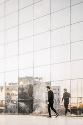 Seitenansicht eines jungen Mannes in voller Länge, der an einer Spiegelung auf einem Glasgebäude vorbeigeht - MASF18359