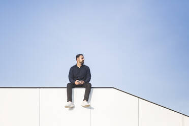 Nachdenklicher Mann, der wegschaut, während er auf einer gebauten Struktur vor blauem Himmel sitzt - MASF18360